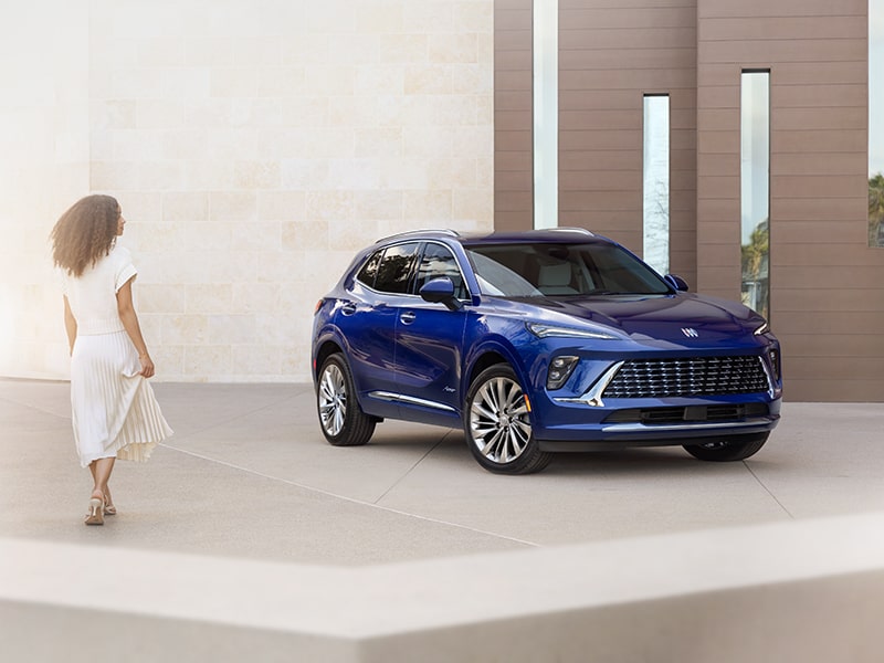 Front-side View of the 2025 Buick Envision Avenir Compact Luxury SUV parked Near a Concrete Building as a person passes by.