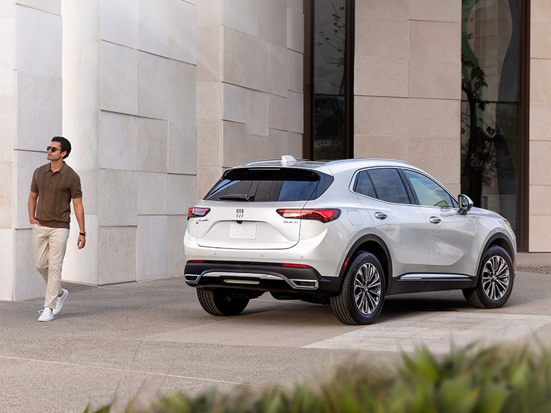 Vue latérale arrière du VUS compact Buick Envision 2025 stationné devant un bâtiment avec une personne qui passe à côté.